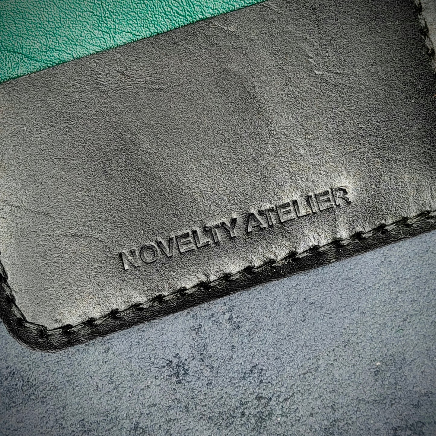 Close up of The Copperfield bifold card holder by Novelty Atelier detailed view of brand name logo embossed into Black and Green full grain leather. Features precise saddle stitching in black thread and hand finished edges. Symbol of artisanal craftsmanship and functional luxury.