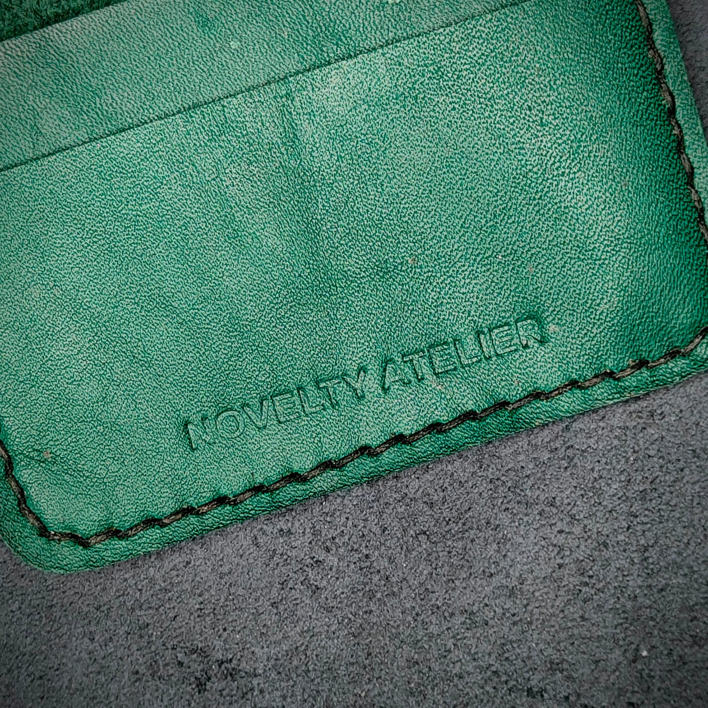 Close up of The Copperfield bifold card holder by Novelty Atelier detailed view of brand name logo embossed into Green full grain leather. Features precise saddle stitching in black thread and hand finished edges. Symbol of artisanal craftsmanship and functional luxury.