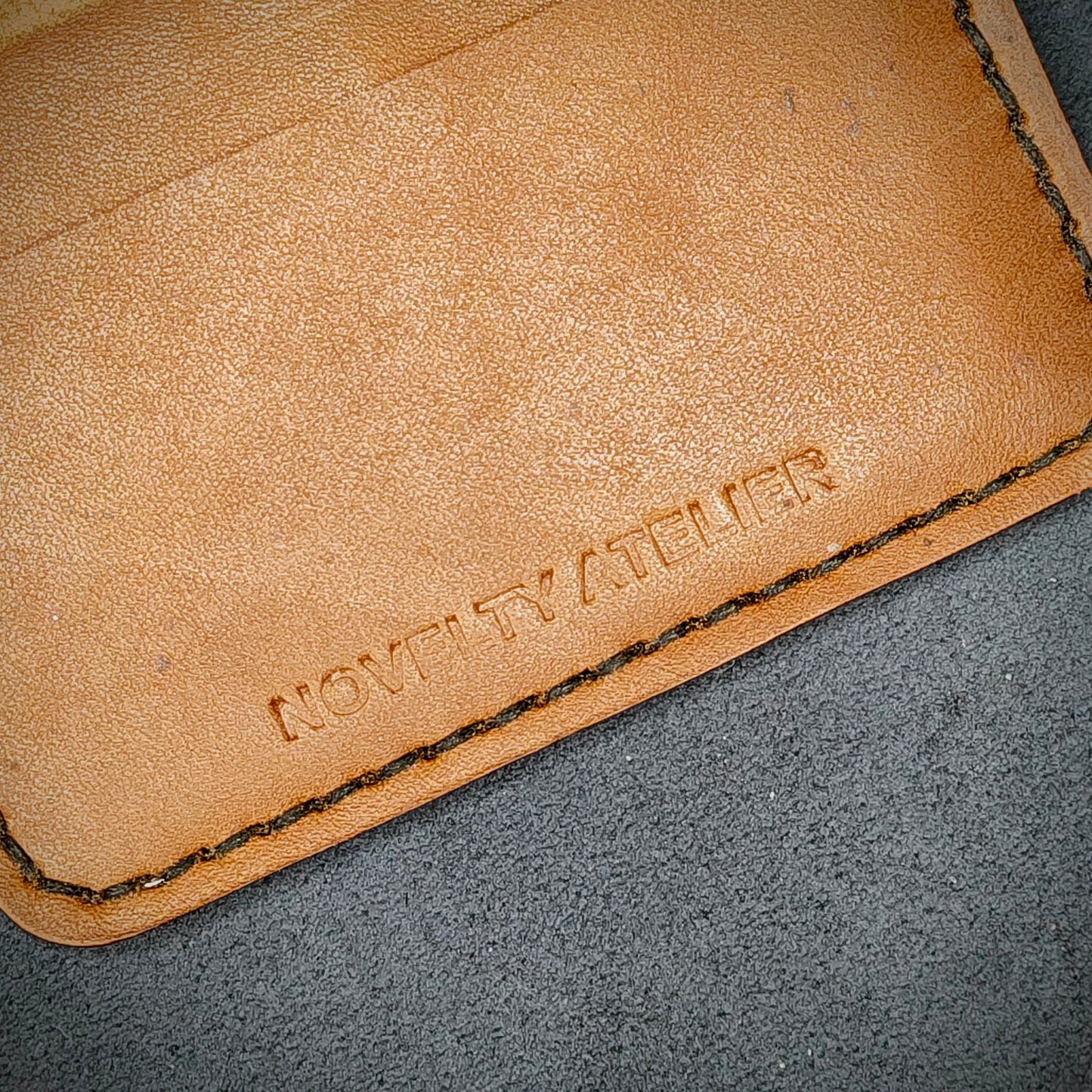 Close up of The Copperfield bifold card holder by Novelty Atelier detailed view of brand name logo embossed into Tann full grain leather. Features precise saddle stitching in black thread and hand finished edges. Symbol of artisanal craftsmanship and functional luxury.