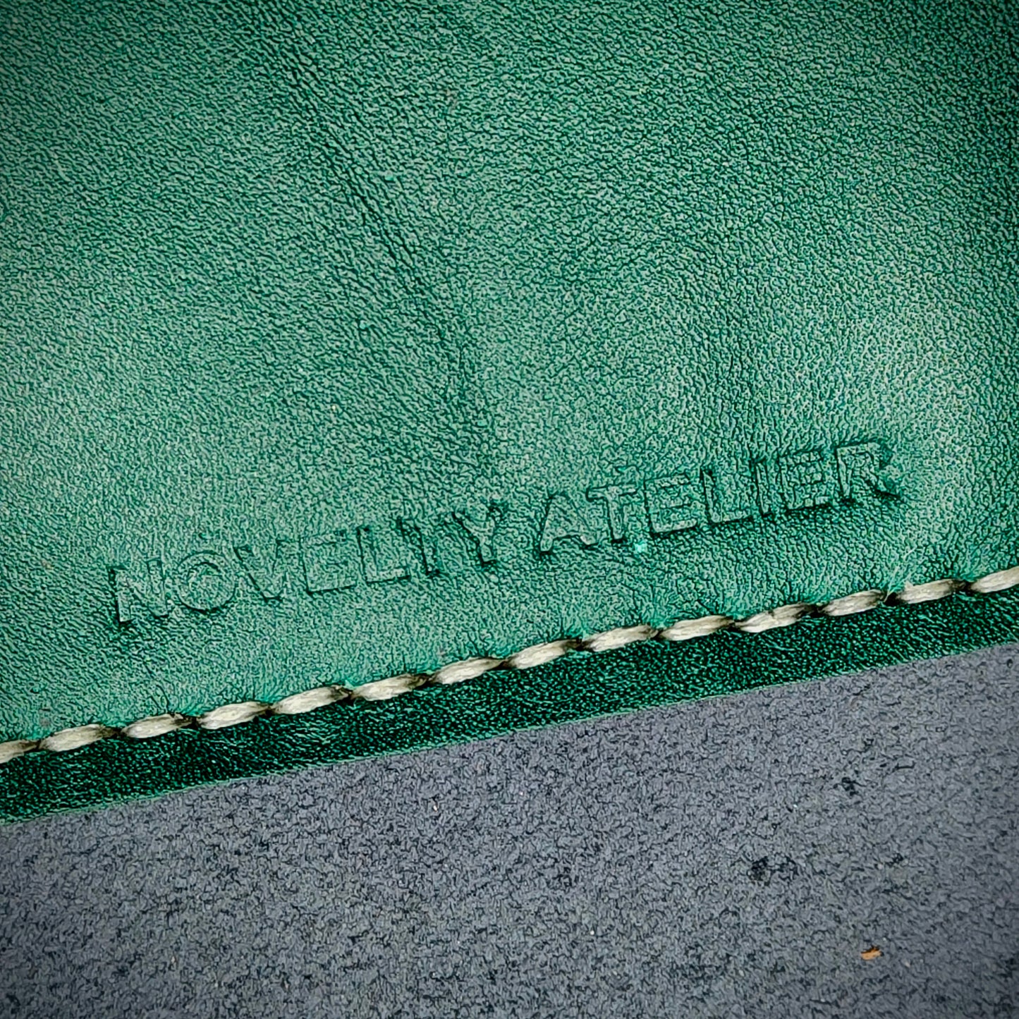 Front view close up shot of the Novelty Atelier brand emboss on The Houdini card holder – detailed close-up of premium black and green full-grain leather and contrast off white hand-stitching . Emphasizes the sharp, elegant logo impression and tactile texture, appealing to luxury EDC and minimalist gear collectors.