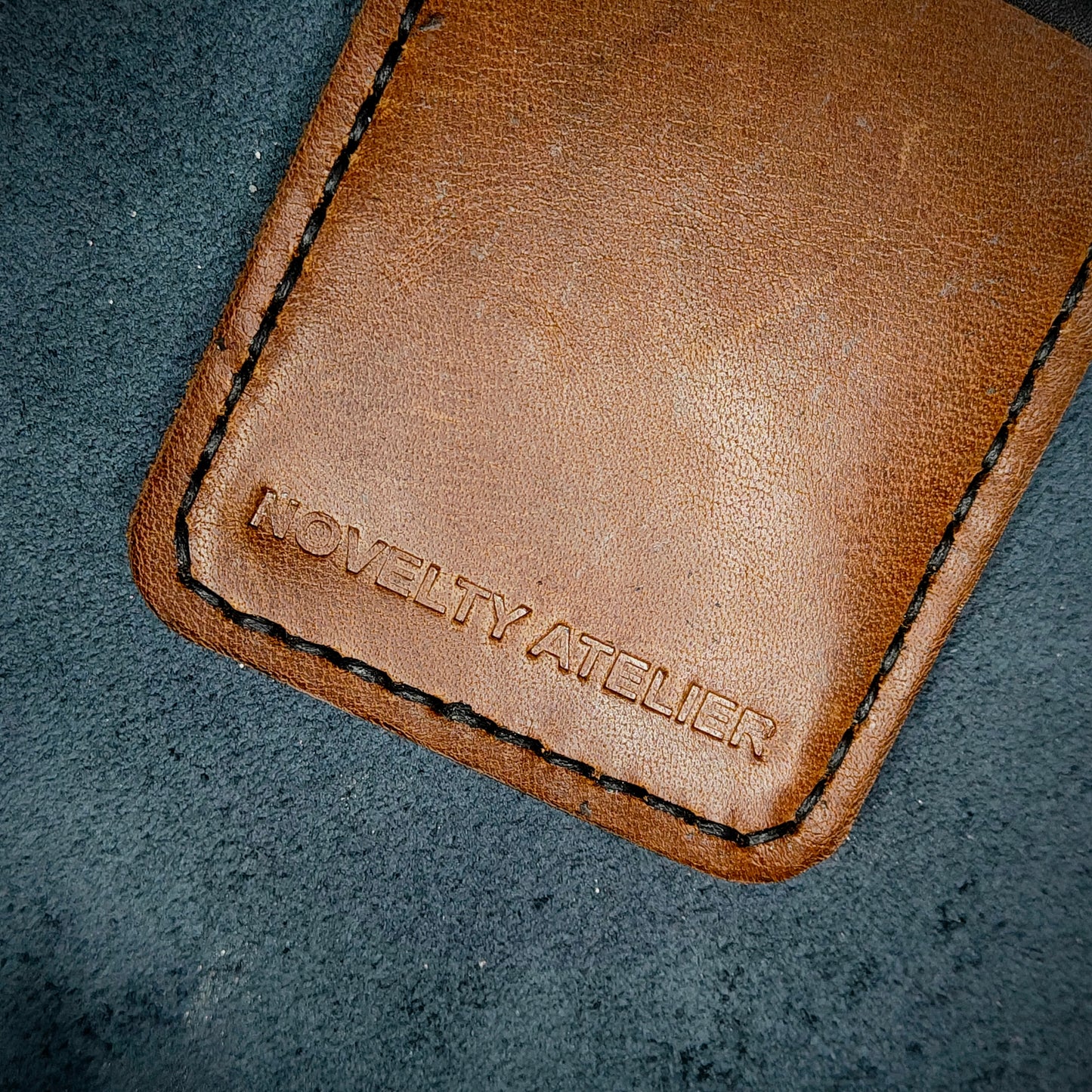 Front view close up shot of the Novelty Atelier brand emboss on The Houdini card holder – detailed close-up of premium Black and Brown full-grain leather and contrast Black hand-stitching . Emphasizes the sharp, elegant logo impression and tactile texture, appealing to luxury EDC and minimalist gear collectors.
