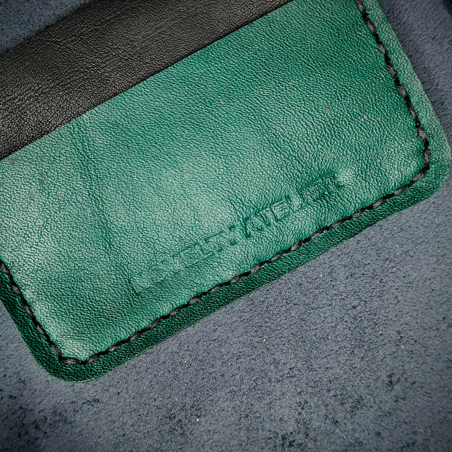 Front view close up shot of the Novelty Atelier brand emboss on The Houdini card holder – detailed close-up of premium black and green full-grain leather and contrast black hand-stitching . Emphasizes the sharp, elegant logo impression and tactile texture, appealing to luxury EDC and minimalist gear collectors.