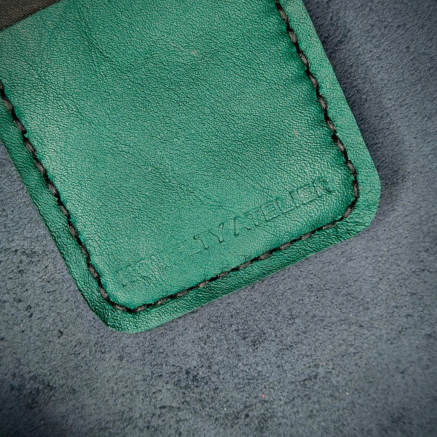 Front view close up shot of the Novelty Atelier brand emboss on The Houdini card holder – detailed close-up of premium black and green full-grain leather and contrast black hand-stitching . Emphasizes the sharp, elegant logo impression and tactile texture, appealing to luxury EDC and minimalist gear collectors.