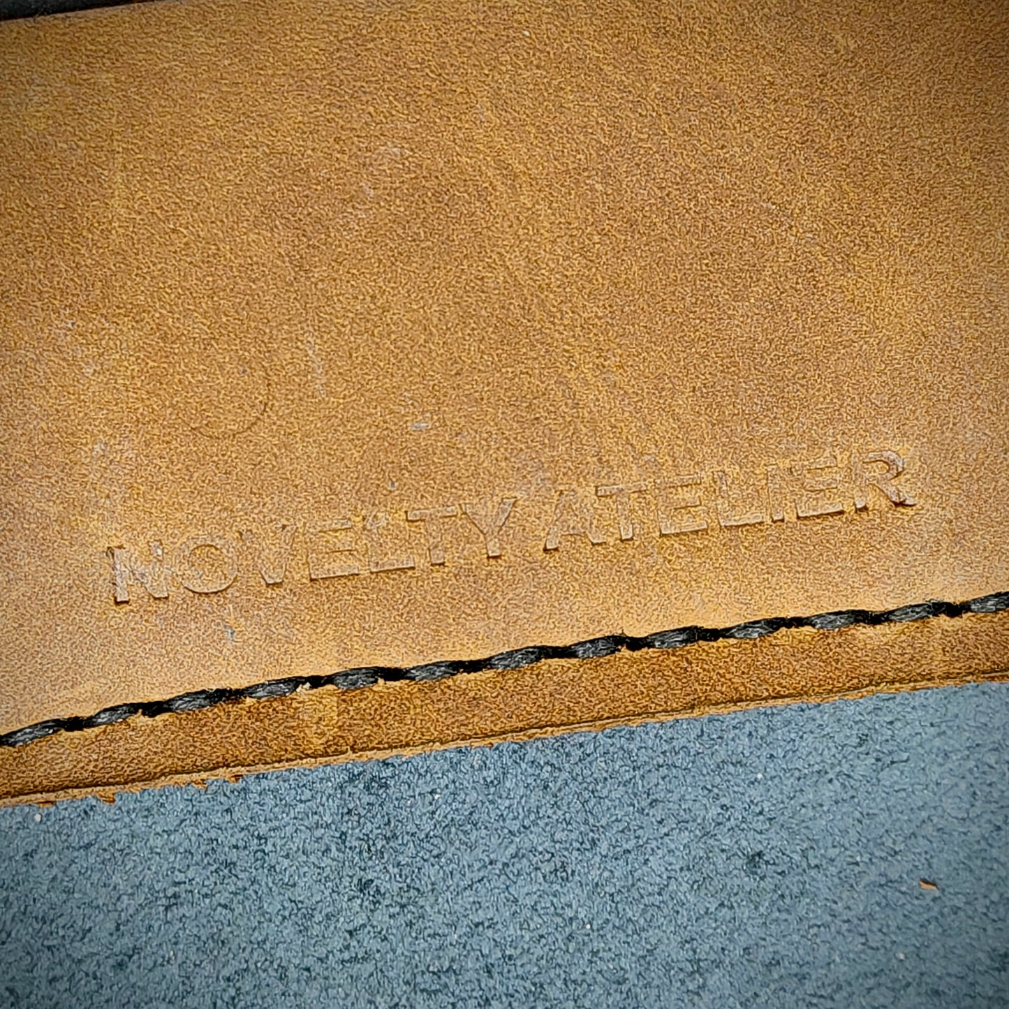Front view close up shot of the Novelty Atelier brand emboss on The Houdini card holder – detailed close-up of premium black and sandstorm Brown full-grain leather and contrast black hand-stitching . Emphasizes the sharp, elegant logo impression and tactile texture, appealing to luxury EDC and minimalist gear collectors.