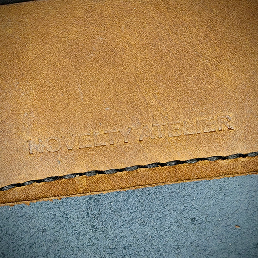 Front view close up shot of the Novelty Atelier brand emboss on The Houdini card holder – detailed close-up of premium black and sandstorm Brown full-grain leather and contrast black hand-stitching . Emphasizes the sharp, elegant logo impression and tactile texture, appealing to luxury EDC and minimalist gear collectors.