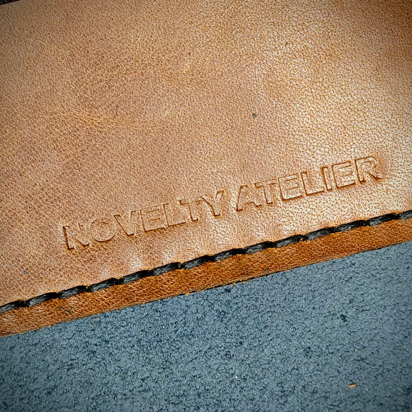 Front view close up shot of the Novelty Atelier brand emboss on The Houdini card holder horizontal configuration– detailed close-up of premium Black and Brown full-grain leather and contrast Black hand-stitching . Emphasizes the sharp, elegant logo impression and tactile texture, appealing to luxury EDC and minimalist gear collectors.