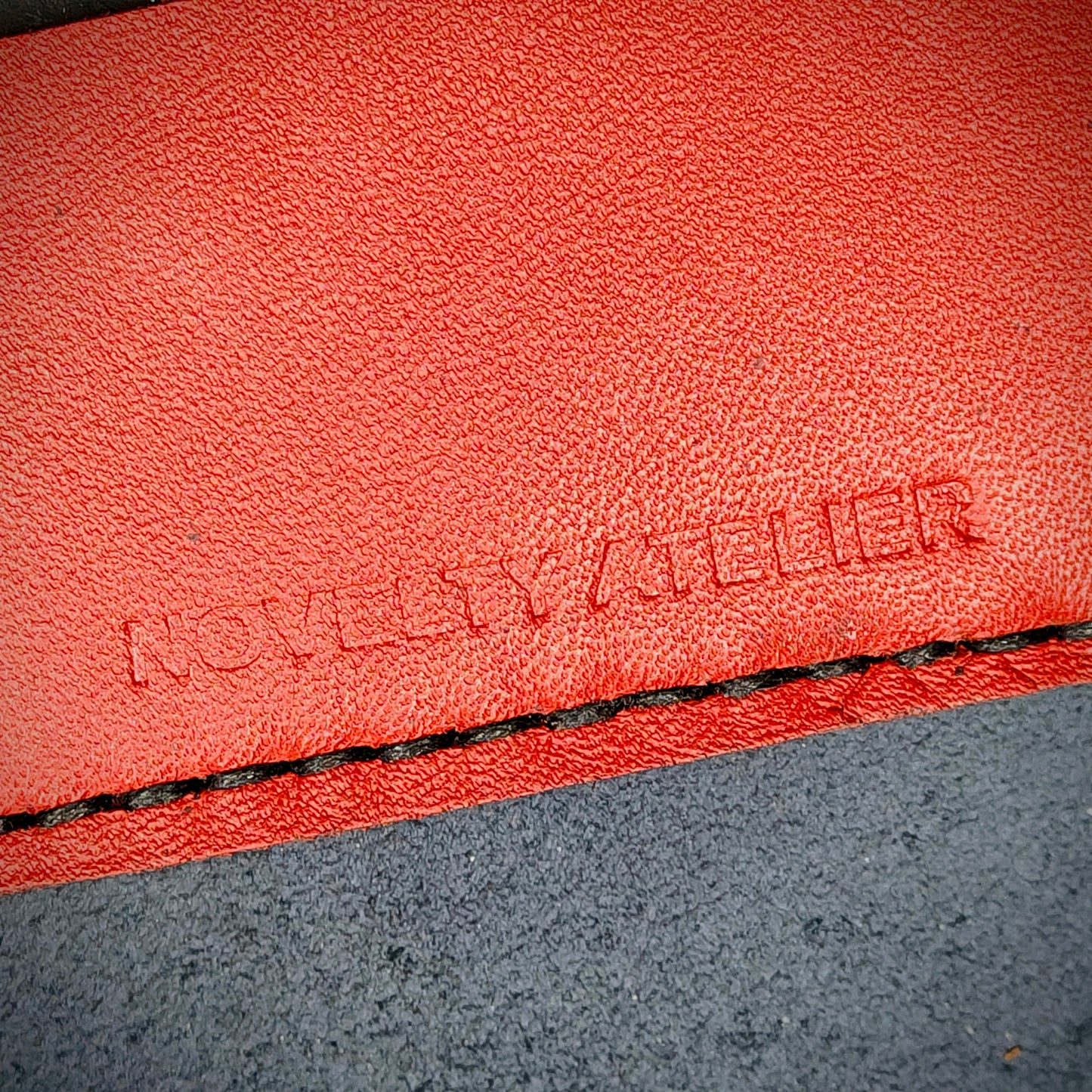 Front view close up shot of the Novelty Atelier brand emboss on The Houdini card holder horizontal configuration– detailed close-up of premium Black and Red full-grain leather and contrast Black hand-stitching . Emphasizes the sharp, elegant logo impression and tactile texture, appealing to luxury EDC and minimalist gear collectors.