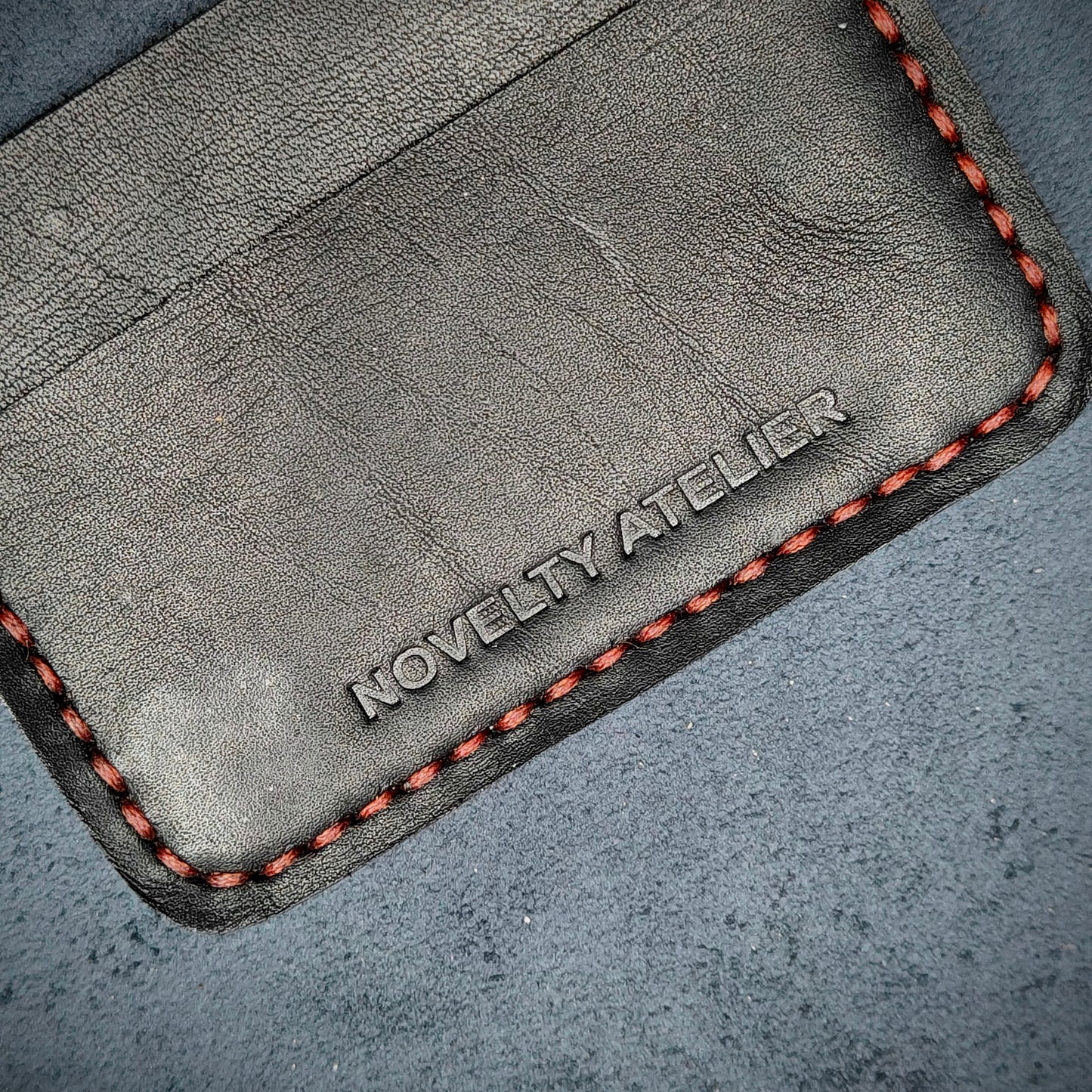Front view close up shot of the Novelty Atelier brand emboss on The Houdini card holder in horizontal configuration– detailed close-up of premium Black full-grain leather and contrast red hand-stitching .Emphasizes the sharp, elegant logo impression and tactile texture, appealing to luxury EDC and minimalist gear collectors.
