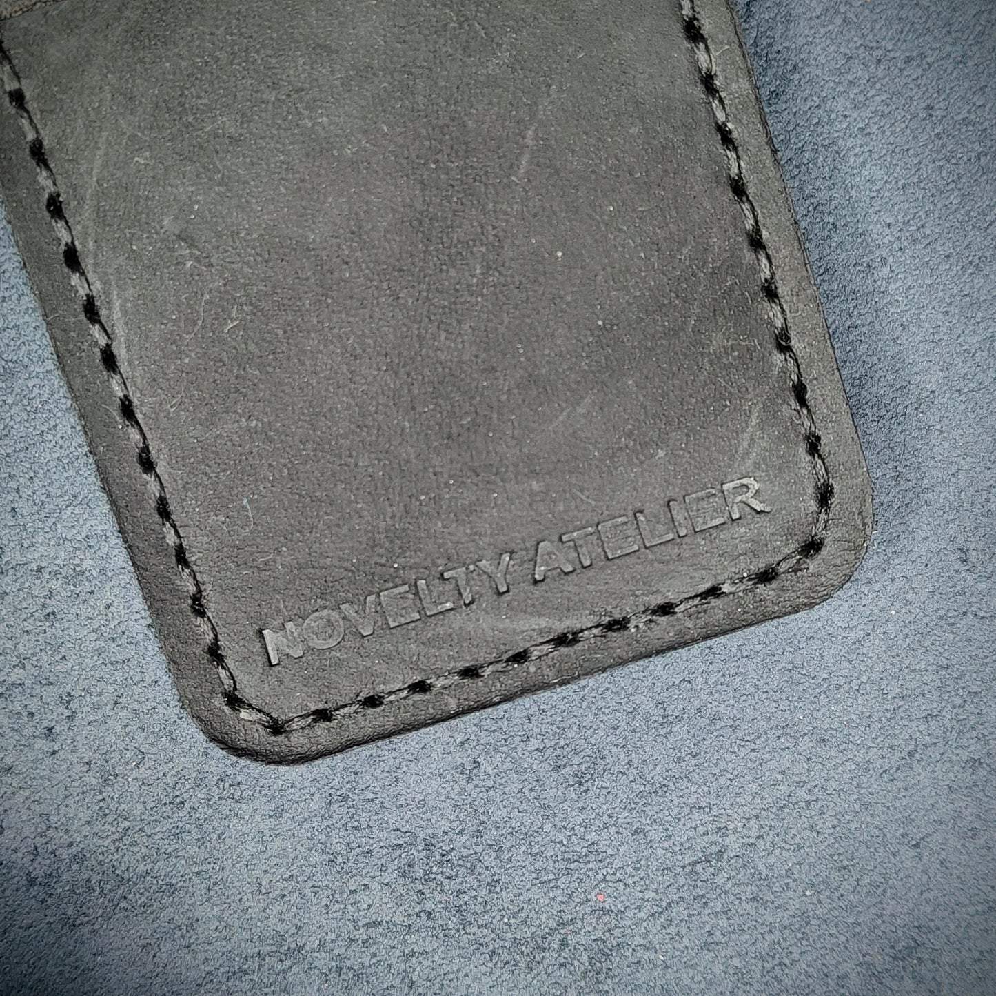 Front view close up shot of the Novelty Atelier brand emboss on The Houdini card holder in vertical configuration– detailed close-up of premium Black full-grain leather and contrast Black hand-stitching . Emphasizes the sharp, elegant logo impression and tactile texture, appealing to luxury EDC and minimalist gear collectors.
