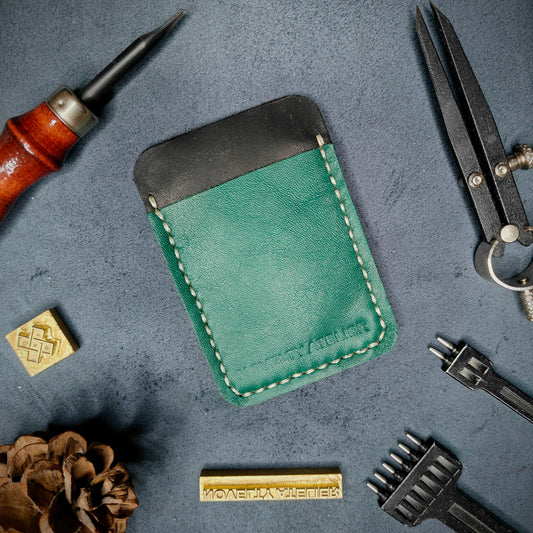 Front view of The Houdini minimalist card holder in vertical configuration by Novelty Atelier – handcrafted from black and green ethically sourced full-grain leather with contrasting off white saddle stitching. Features the “Novelty Atelier” brand name cleanly embossed on the front. Perfectly balanced minimalist wallet design tailored for modern EDC enthusiasts who value craftsmanship and brand integrity.