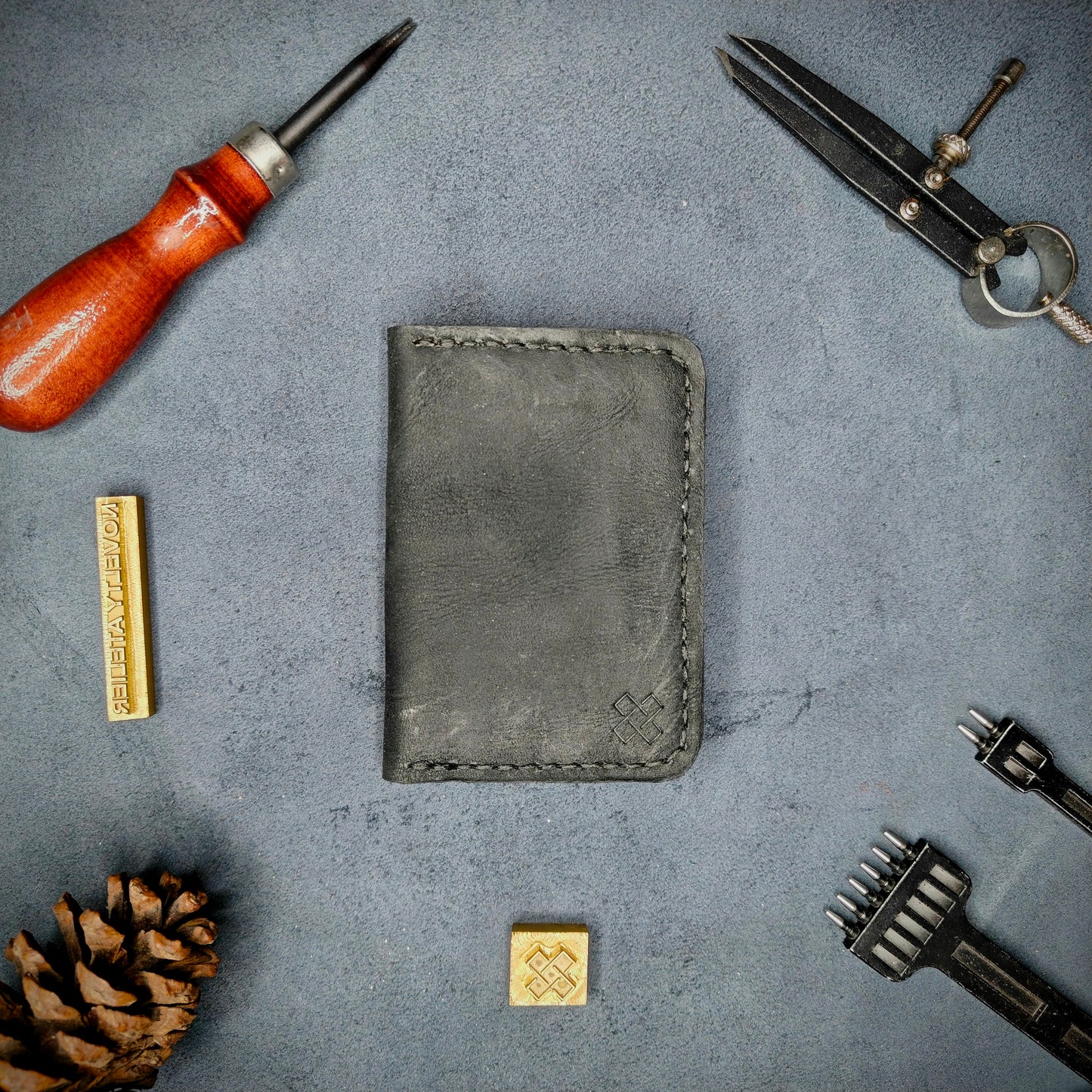 The Copperfield full grain leather bifold wallet by Novelty Atelier closed view highlighting slim and rugged design. Handcrafted from Black and Brown ethically sourced leather, saddle stitched in black thread. Set against a textured background with branding tools and pine cone, perfect for minimalist EDC style.