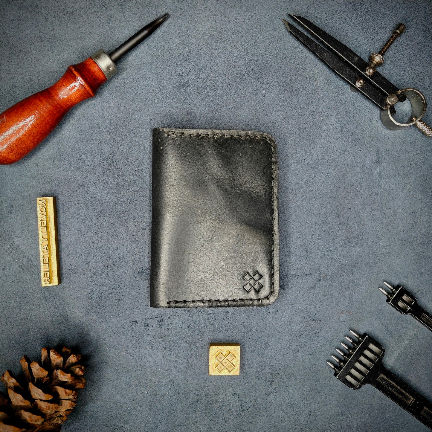 The Copperfield full grain leather bifold wallet by Novelty Atelier closed view highlighting slim and rugged design. Handcrafted from Black and Green ethically sourced leather, saddle stitched in black thread. Set against a textured background with branding tools and pine cone, perfect for minimalist EDC style.