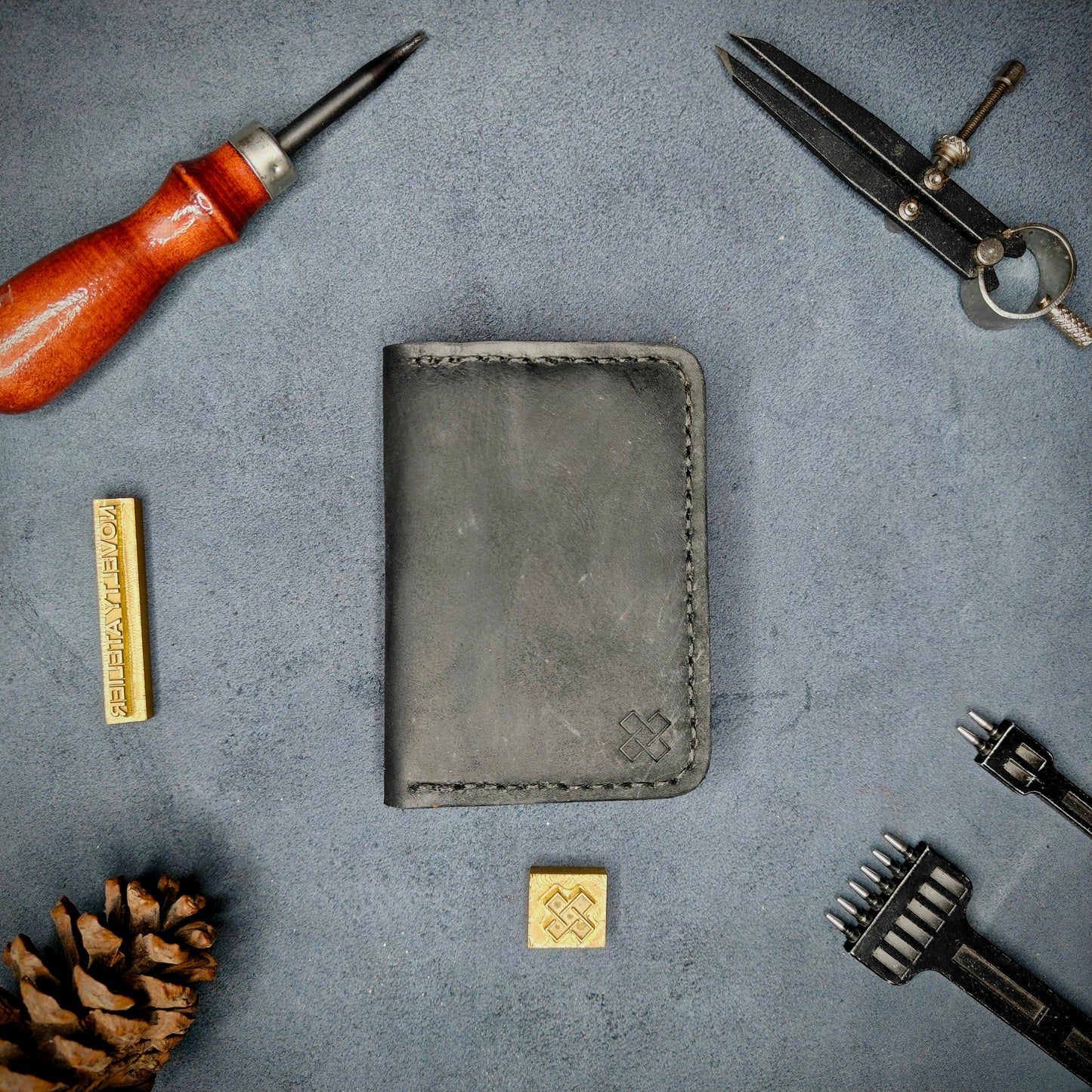 The Copperfield full grain leather bifold wallet by Novelty Atelier closed view highlighting slim and rugged design. Handcrafted from Black and Mustard yellow ethically sourced leather, saddle stitched in black thread. Set against a textured background with branding tools and pine cone, perfect for minimalist EDC style.
