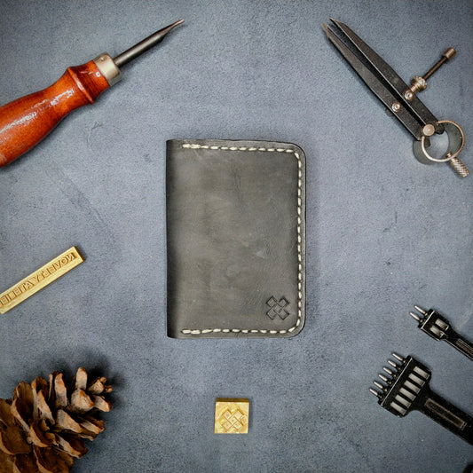 The Copperfield full grain leather bifold wallet by Novelty Atelier closed view highlighting slim and rugged design. Handcrafted from Black and Mustard yellow ethically sourced leather, saddle stitched in off white thread. Set against a textured background with branding tools and pine cone, perfect for minimalist EDC style.