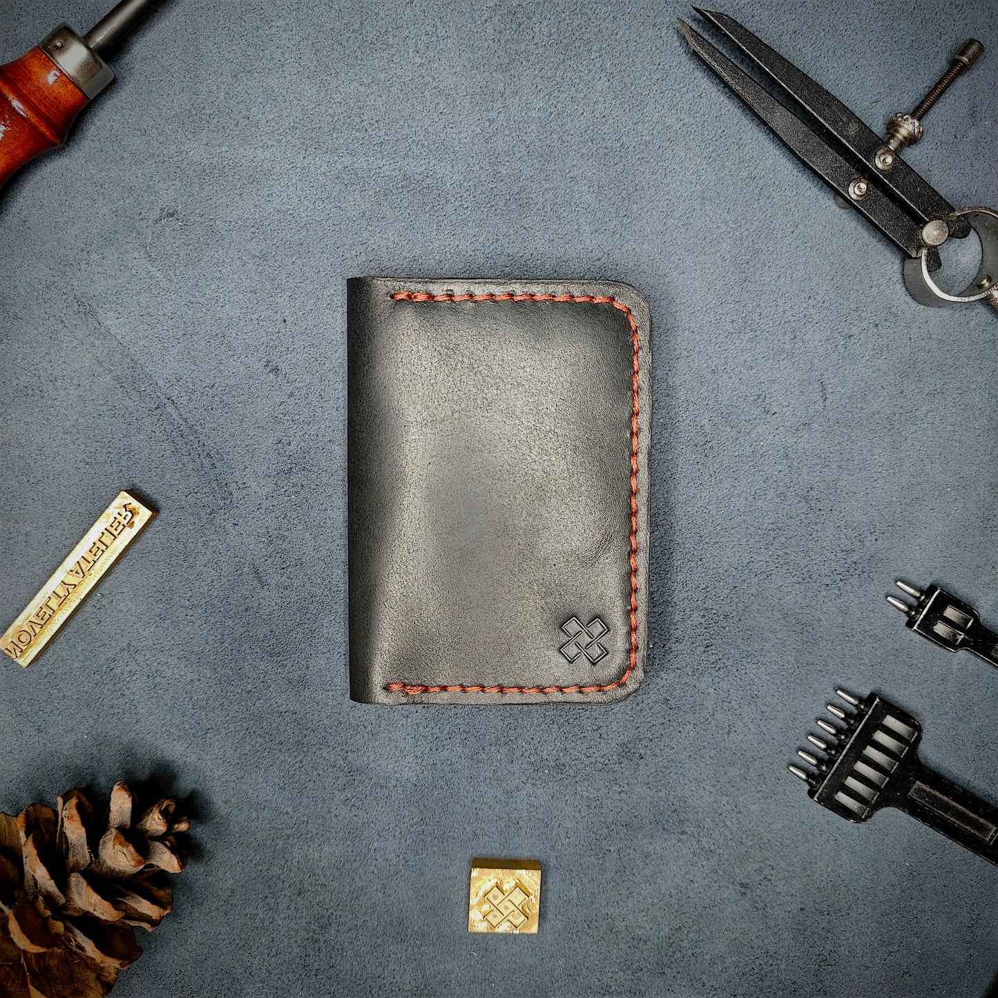 The Copperfield full grain leather bifold wallet by Novelty Atelier closed view highlighting slim and rugged design. Handcrafted from Black ethically sourced leather, saddle stitched in Red thread. Set against a textured background with branding tools and pine cone, perfect for minimalist EDC style.