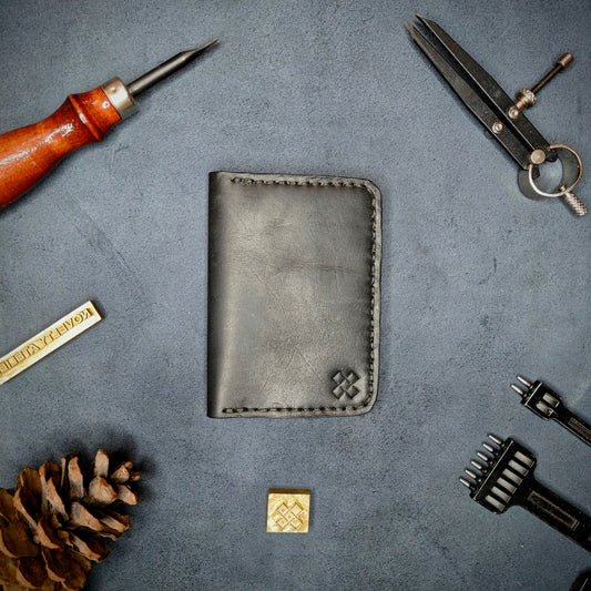 The Copperfield full grain leather bifold wallet by Novelty Atelier closed view highlighting slim and rugged design. Handcrafted from Black ethically sourced leather, saddle stitched in black thread. Set against a textured background with branding tools and pine cone, perfect for minimalist EDC style