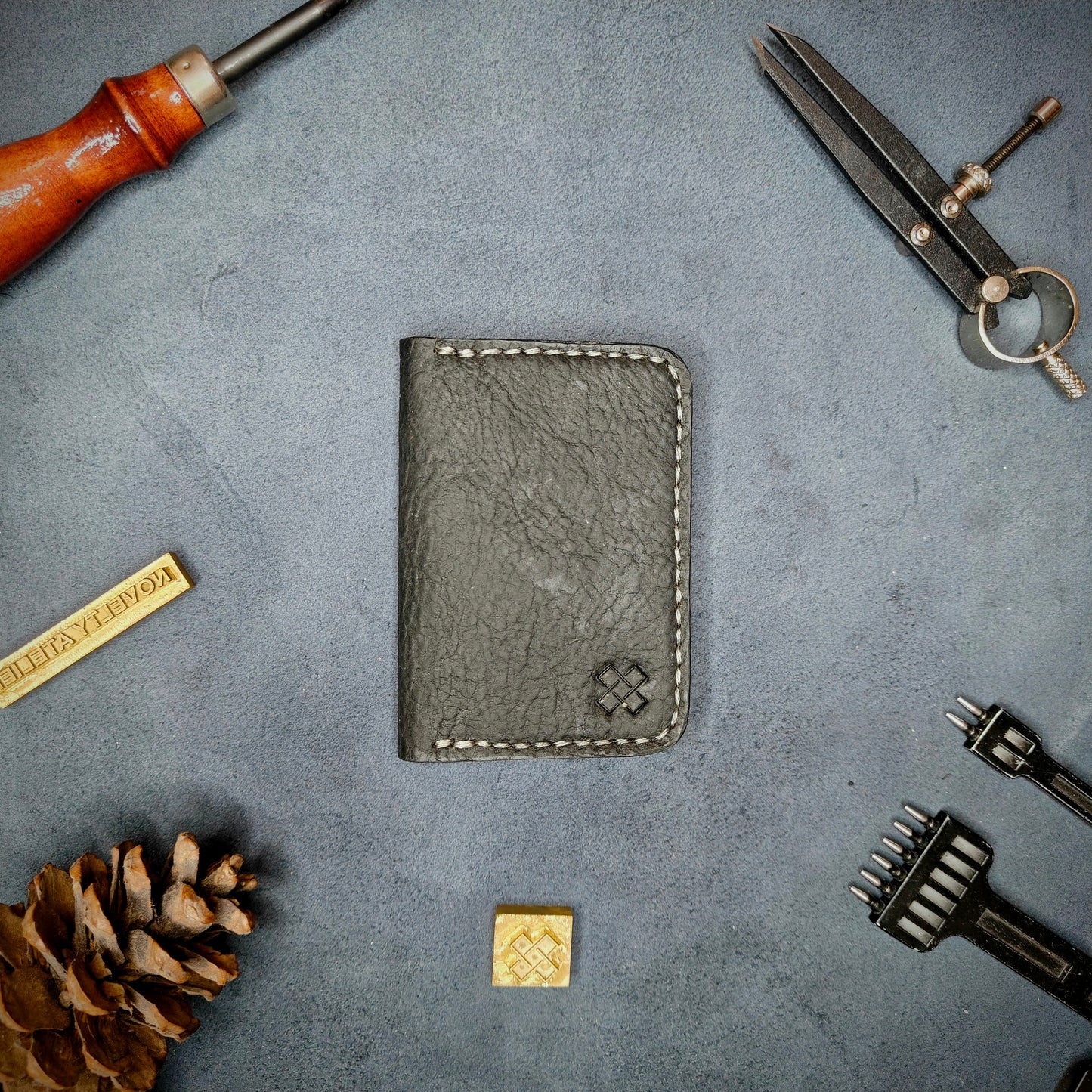 The Copperfield full grain leather bifold wallet by Novelty Atelier closed view highlighting slim and rugged design. Handcrafted from Black ethically sourced leather, saddle stitched in off white thread. Set against a textured background with branding tools and pine cone, perfect for minimalist EDC style.