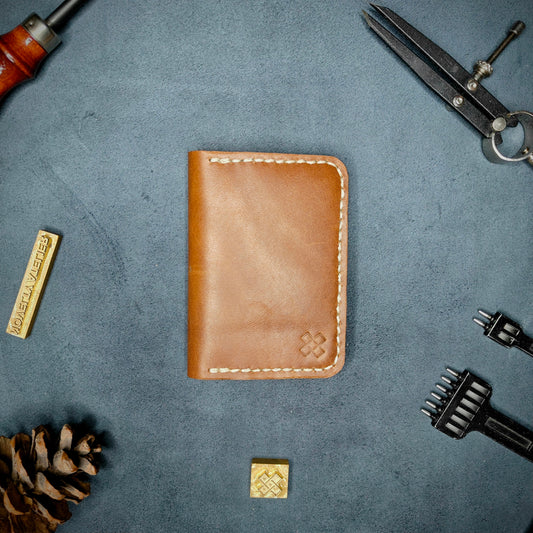 The Copperfield full grain leather bifold wallet by Novelty Atelier closed view highlighting slim and rugged design. Handcrafted from Brown ethically sourced leather, saddle stitched in off white thread. Set against a textured background with branding tools and pine cone, perfect for minimalist EDC style.