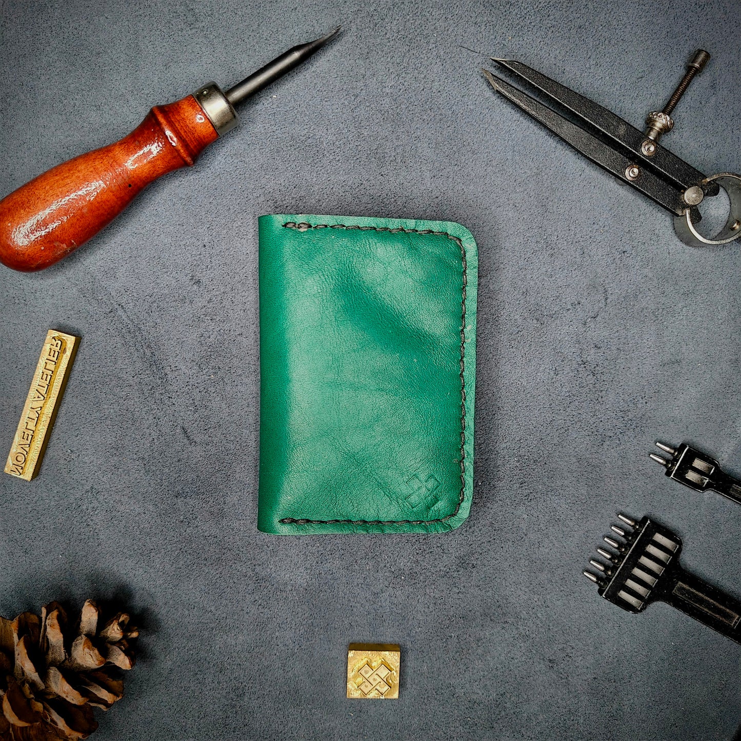 The Copperfield full grain leather bifold wallet by Novelty Atelier closed view highlighting slim and rugged design. Handcrafted from Green ethically sourced leather, saddle stitched in black thread. Set against a textured background with branding tools and pine cone, perfect for minimalist EDC style.