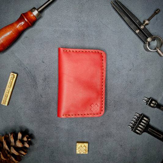 The Copperfield full grain leather bifold wallet by Novelty Atelier closed view highlighting slim and rugged design. Handcrafted from Red ethically sourced leather, saddle stitched in Red thread. Set against a textured background with branding tools and pine cone, perfect for minimalist EDC style.