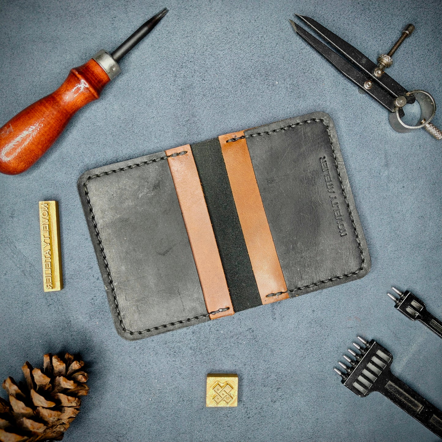 The Copperfield handmade leather card holder by Novelty Atelier open flat view revealing 4 card slots, crafted from ethically sourced Black and Brown full grain leather with black thread saddle stitching. Displayed on suede with brass stamp, leatherworking tools, and pine cone for EDC inspired composition. A durable, minimalist wallet for everyday carry.