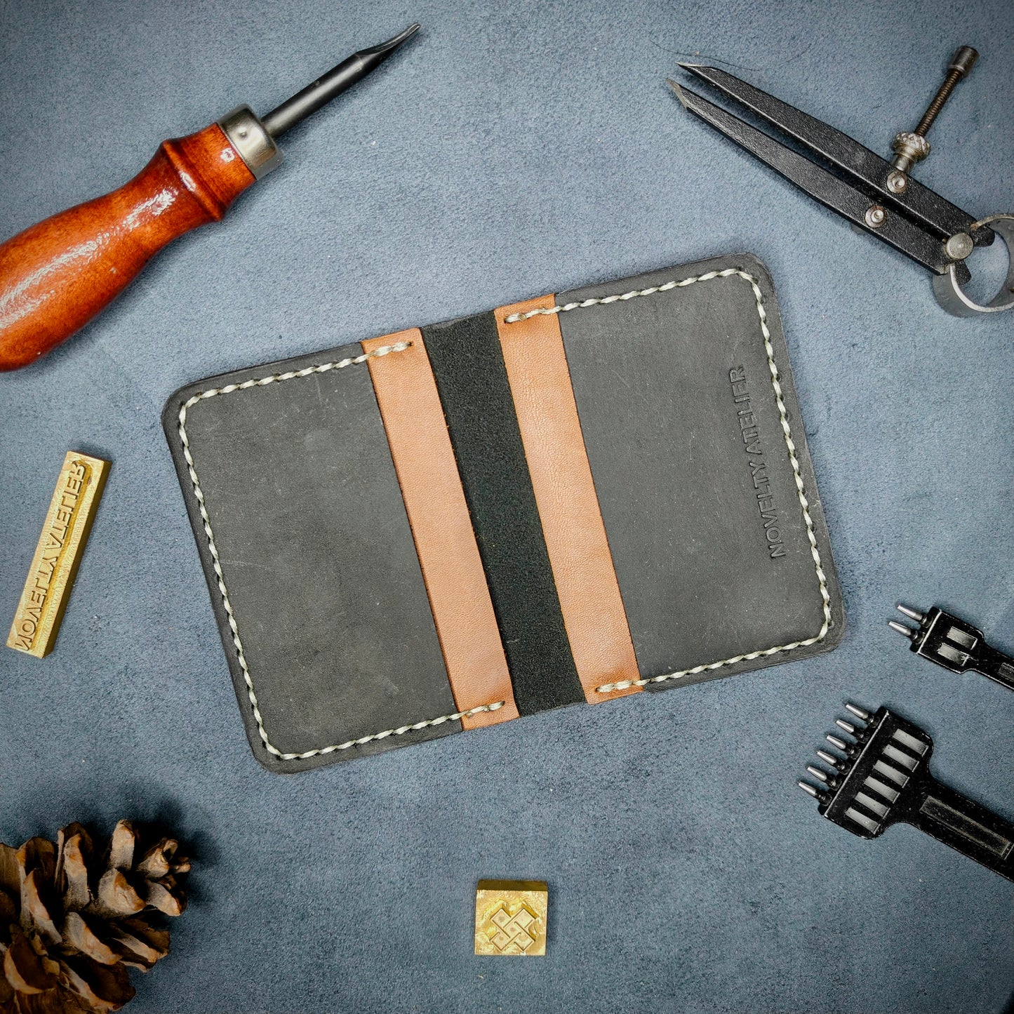 The Copperfield handmade leather card holder by Novelty Atelier open flat view revealing 4 card slots, crafted from ethically sourced Black and Brown full grain leather with off white saddle stitching. Displayed on suede with brass stamp, leatherworking tools, and pine cone for EDC inspired composition. A durable, minimalist wallet for everyday carry.