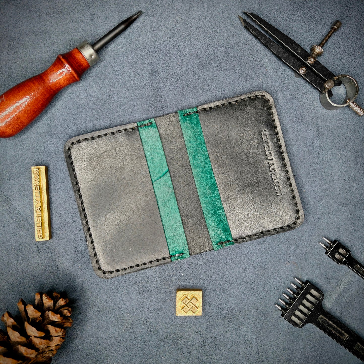 The Copperfield handmade leather card holder by Novelty Atelier open flat view revealing 4 card slots, crafted from ethically sourced Black and Green full grain leather with black thread saddle stitching. Displayed on suede with brass stamp, leatherworking tools, and pine cone for EDC inspired composition. A durable, minimalist wallet for everyday carry.