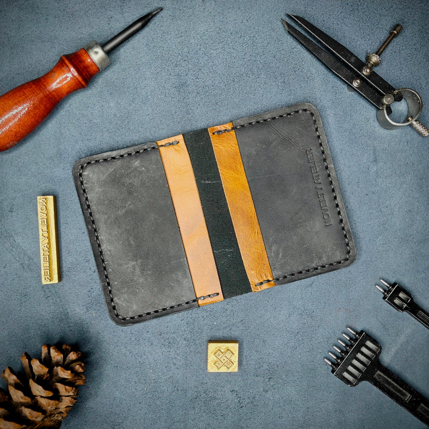 The Copperfield handmade leather card holder by Novelty Atelier open flat view revealing 4 card slots, crafted from ethically sourced Black and Mustard full grain leather with black thread saddle stitching. Displayed on suede with brass stamp, leatherworking tools, and pine cone for EDC inspired composition. A durable, minimalist wallet for everyday carry.