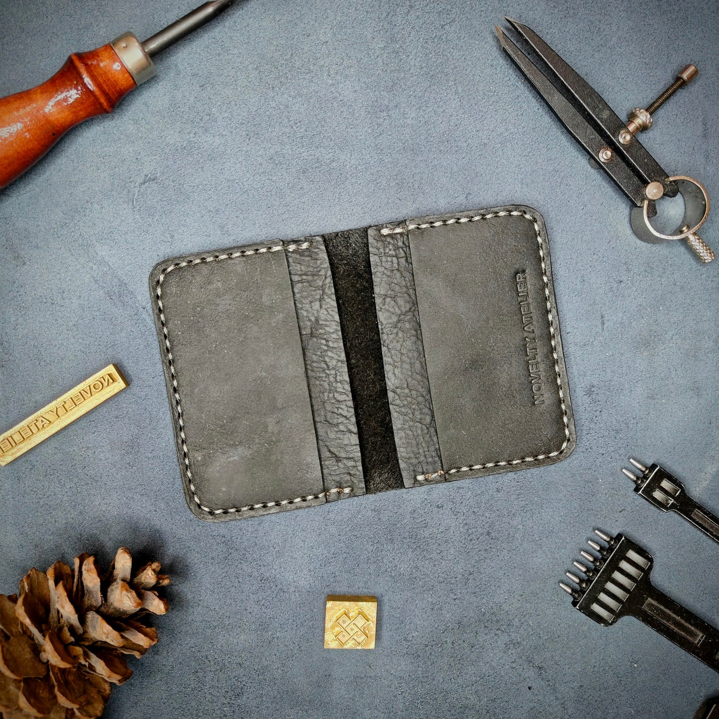 The Copperfield handmade leather card holder by Novelty Atelier open flat view revealing 4 card slots, crafted from ethically sourced Black full grain leather with off white saddle stitching. Displayed on suede with brass stamp, leatherworking tools, and pine cone for EDC inspired composition. A durable, minimalist wallet for everyday carry.