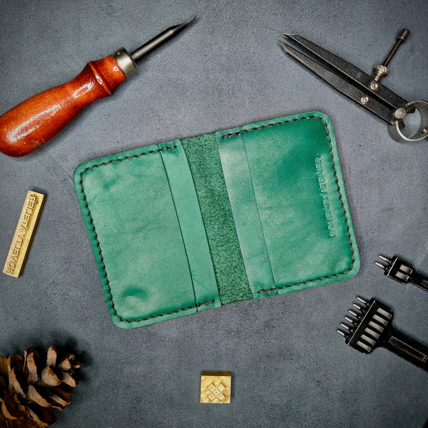 The Copperfield handmade leather card holder by Novelty Atelier open flat view revealing 4 card slots, crafted from ethically sourced Green full grain leather with black thread saddle stitching. Displayed on suede with brass stamp, leatherworking tools, and pine cone for EDC inspired composition. A durable, minimalist wallet for everyday carry.