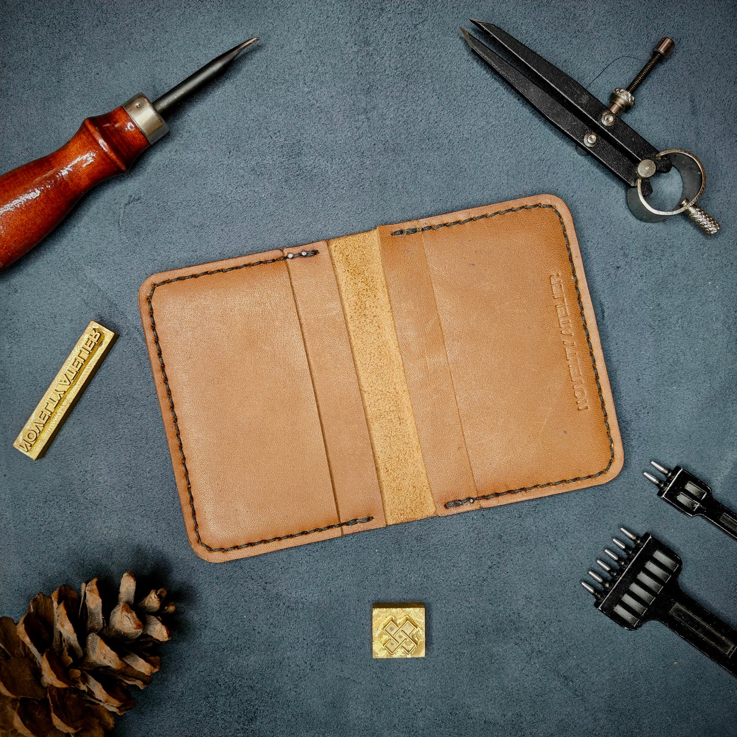 The Copperfield handmade leather card holder by Novelty Atelier open flat view revealing 4 card slots, crafted from ethically sourced Tann full grain leather with black thread saddle stitching. Displayed on suede with brass stamp, leatherworking tools, and pine cone for EDC inspired composition. A durable, minimalist wallet for everyday carry.