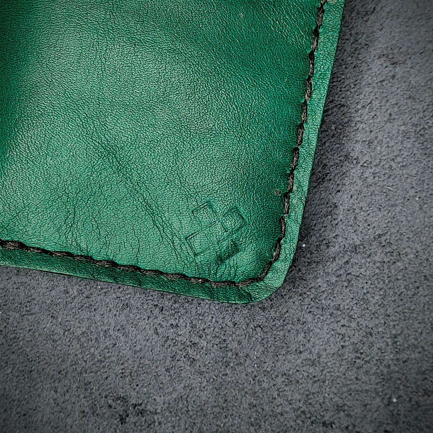Zoomed in view of Novelty Atelier’s signature insignia embossed on The Copperfield minimalist leather card holder. Hand stitched in black thread using traditional saddle technique. Crafted from Green full grain leather, symbolizing timeless durability and ethical sophistication.