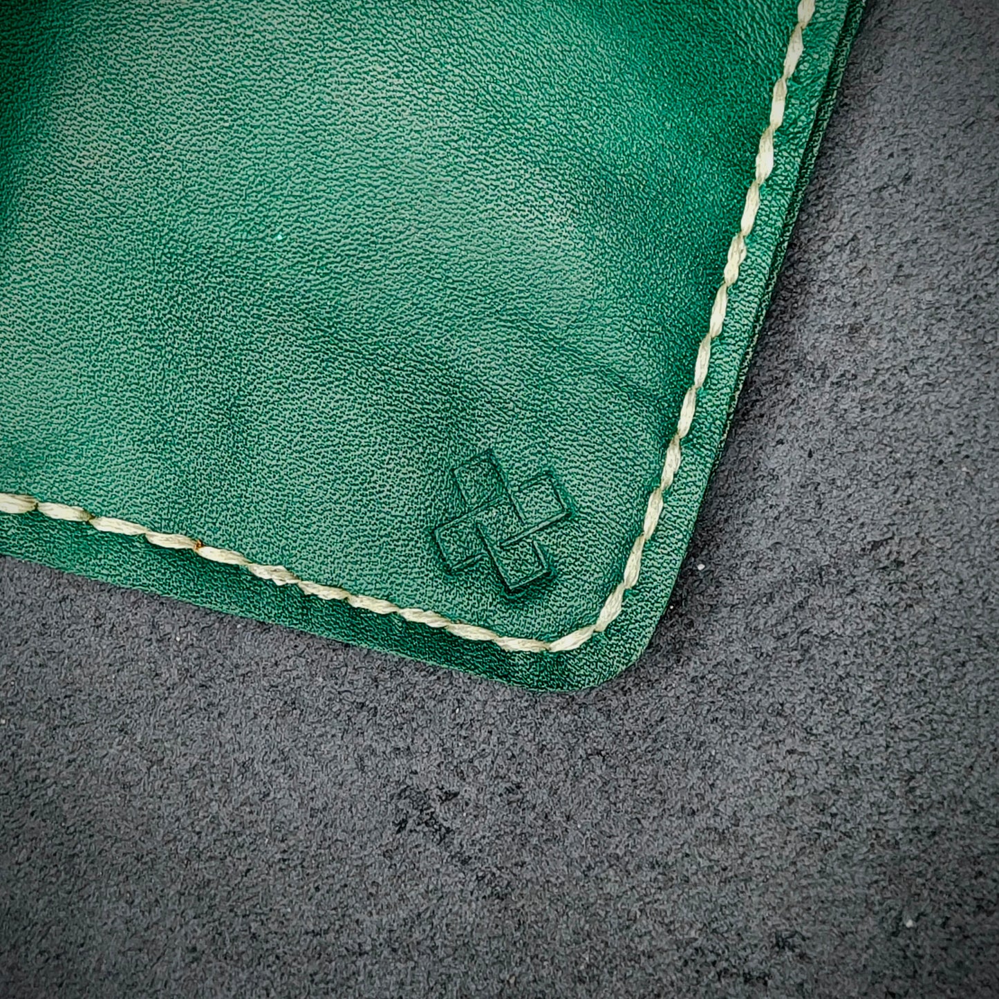 Zoomed in view of Novelty Atelier’s signature insignia embossed on The Copperfield minimalist leather card holder. Hand stitched in off white thread using traditional saddle technique. Crafted from Green full grain leather, symbolizing timeless durability and ethical sophistication.