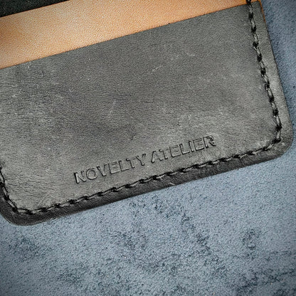 Close up of The Copperfield bifold card holder by Novelty Atelier detailed view of brand name logo embossed into Black and Brown full grain leather. Features precise saddle stitching in black thread and hand finished edges. Symbol of artisanal craftsmanship and functional luxury.
