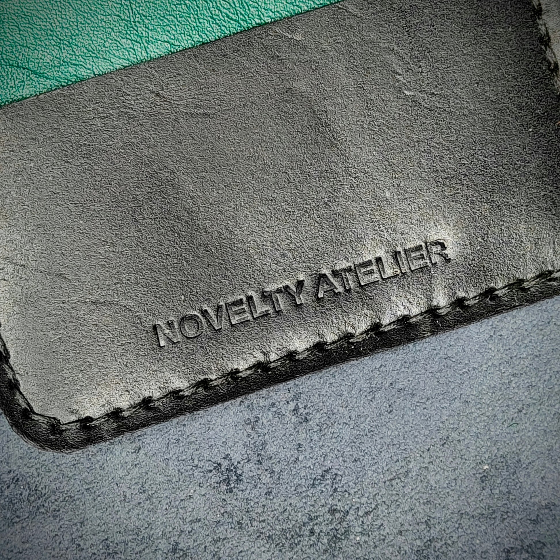 Close up of The Copperfield bifold card holder by Novelty Atelier detailed view of brand name logo embossed into Black and Green full grain leather. Features precise saddle stitching in black thread and hand finished edges. Symbol of artisanal craftsmanship and functional luxury.
