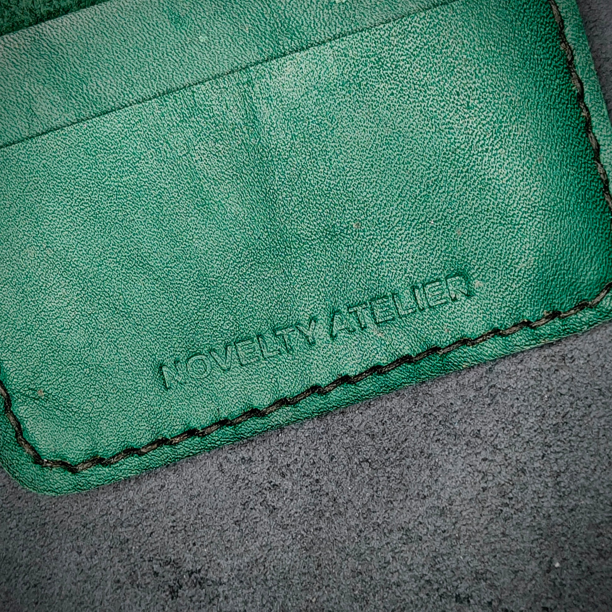 Close up of The Copperfield bifold card holder by Novelty Atelier detailed view of brand name logo embossed into Green full grain leather. Features precise saddle stitching in black thread and hand finished edges. Symbol of artisanal craftsmanship and functional luxury.
