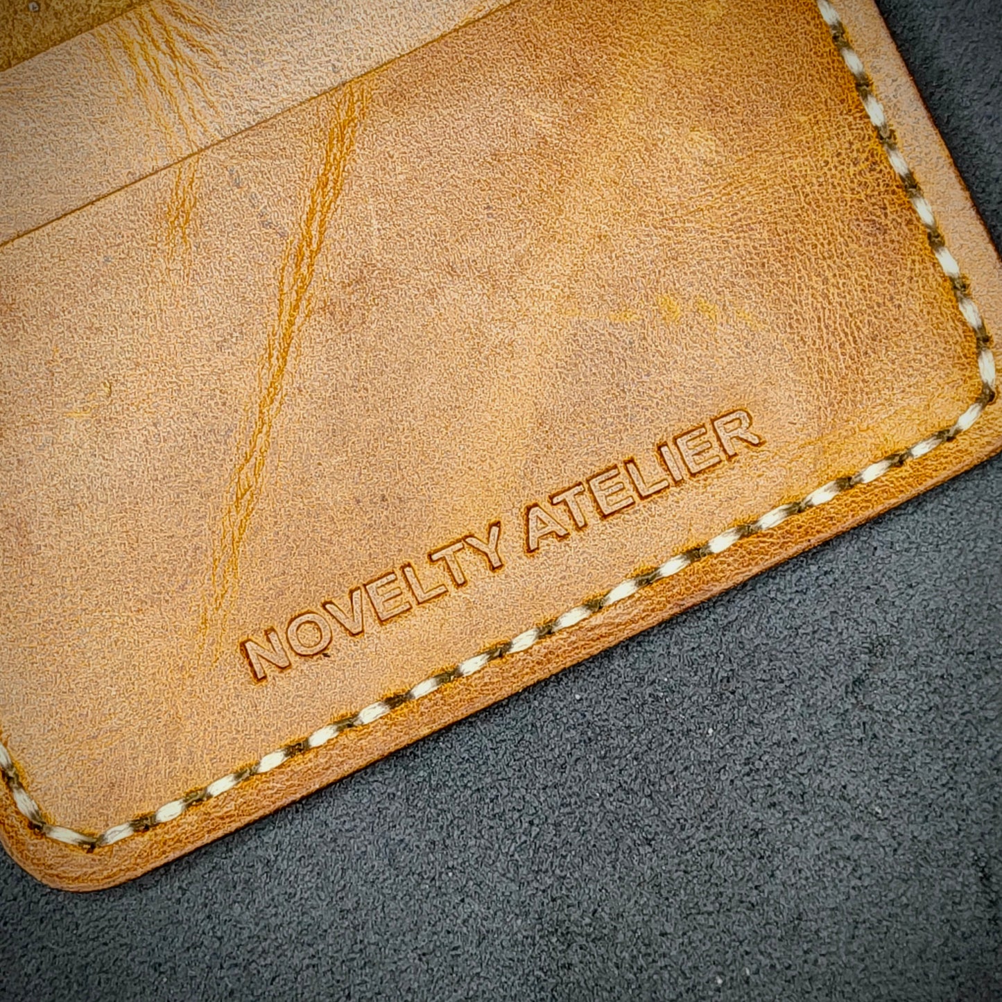 Close up of The Copperfield bifold card holder by Novelty Atelier detailed view of brand name logo embossed into Mustard yellow full grain leather. Features precise saddle stitching in off white  thread and hand finished edges. Symbol of artisanal craftsmanship and functional luxury.
