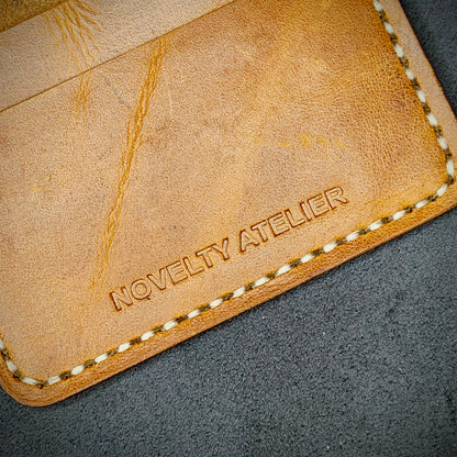 Close up of The Copperfield bifold card holder by Novelty Atelier detailed view of brand name logo embossed into Mustard yellow full grain leather. Features precise saddle stitching in off white  thread and hand finished edges. Symbol of artisanal craftsmanship and functional luxury.
