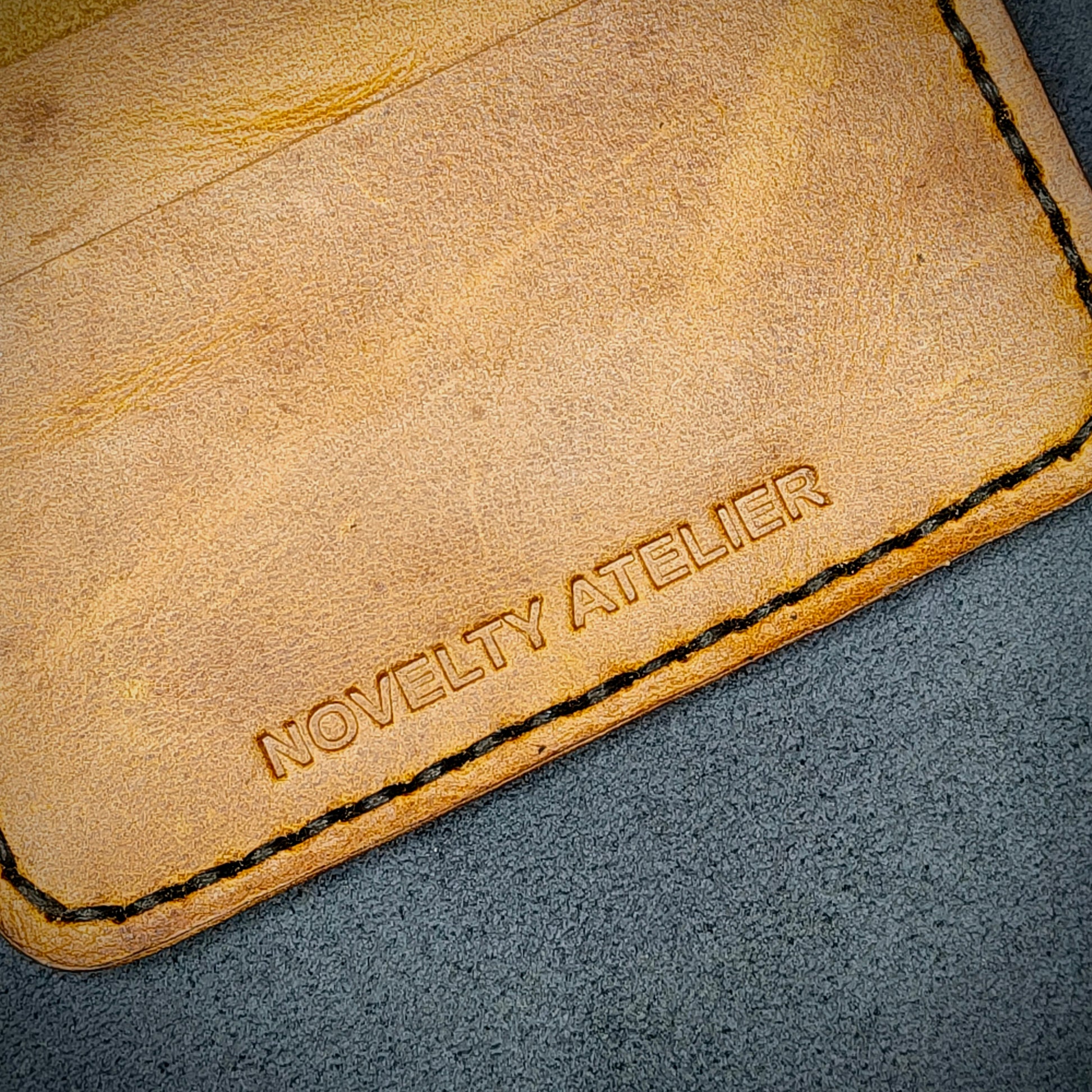 Close up of The Copperfield bifold card holder by Novelty Atelier detailed view of brand name logo embossed into Mustard yellow full grain leather. Features precise saddle stitching in black thread and hand finished edges. Symbol of artisanal craftsmanship and functional luxury.
