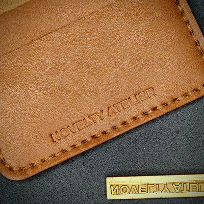 Close up of The Copperfield bifold card holder by Novelty Atelier detailed view of brand name logo embossed into Tann full grain leather. Features precise saddle stitching in Brown thread and hand finished edges. Symbol of artisanal craftsmanship and functional luxury.
