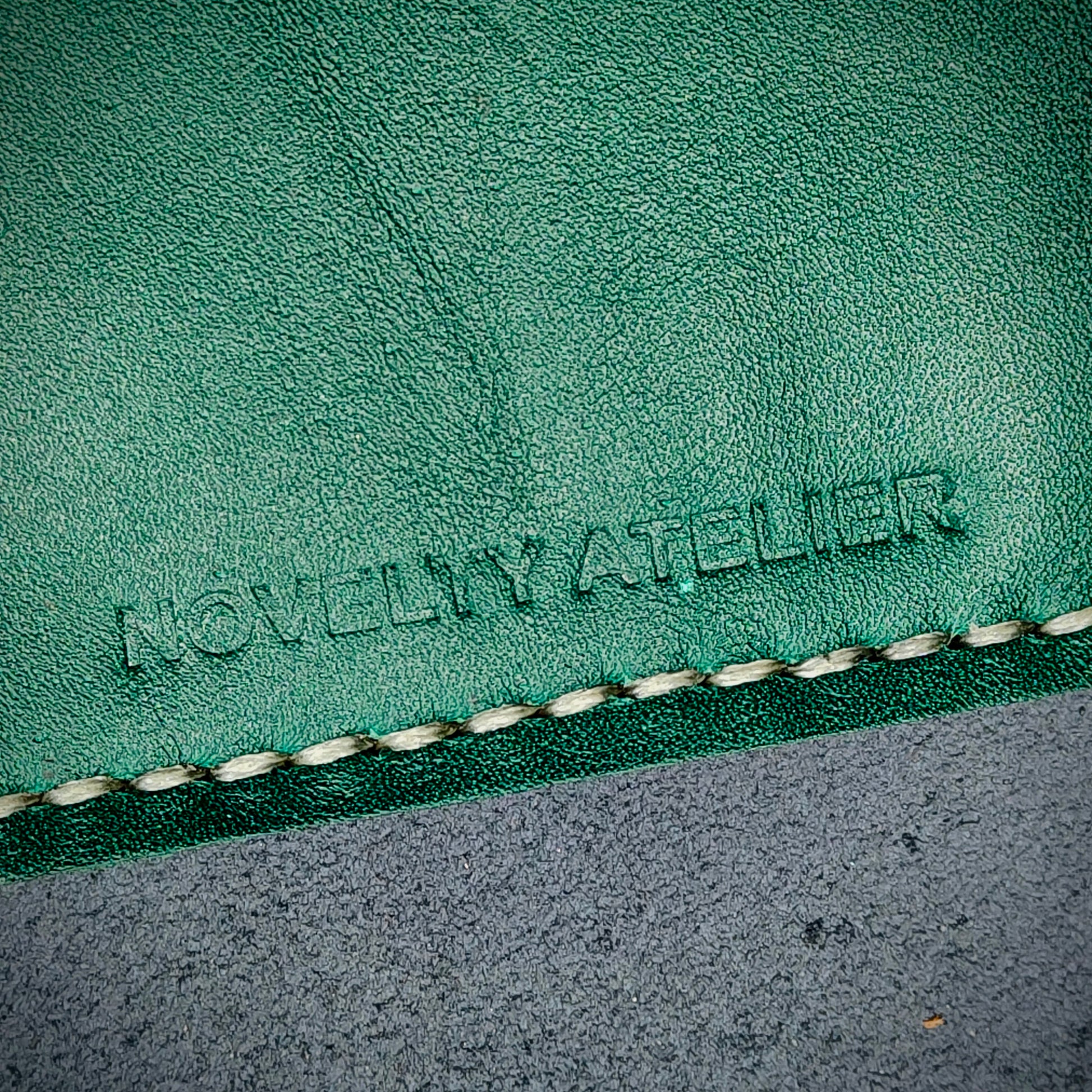 Front view close up shot of the Novelty Atelier brand emboss on The Houdini card holder – detailed close-up of premium black and green full-grain leather and contrast off white hand-stitching . Emphasizes the sharp, elegant logo impression and tactile texture, appealing to luxury EDC and minimalist gear collectors.
