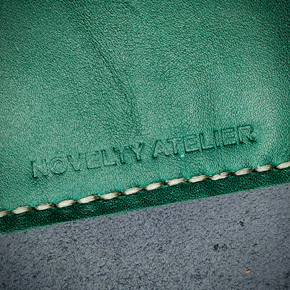 Front view close up shot of the Novelty Atelier brand emboss on The Houdini card holder – detailed close-up of premium black and green full-grain leather and contrast off white hand-stitching . Emphasizes the sharp, elegant logo impression and tactile texture, appealing to luxury EDC and minimalist gear collectors.
