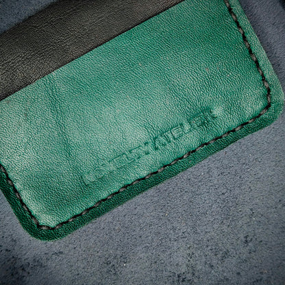 Front view close up shot of the Novelty Atelier brand emboss on The Houdini card holder – detailed close-up of premium black and green full-grain leather and contrast black hand-stitching . Emphasizes the sharp, elegant logo impression and tactile texture, appealing to luxury EDC and minimalist gear collectors.
