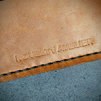 Front view close up shot of the Novelty Atelier brand emboss on The Houdini card holder horizontal configuration– detailed close-up of premium Black and Brown full-grain leather and contrast Black hand-stitching . Emphasizes the sharp, elegant logo impression and tactile texture, appealing to luxury EDC and minimalist gear collectors.
