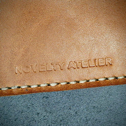 Front view close up shot of the Novelty Atelier brand emboss on The Houdini card holder  horizontal configuration– detailed close-up of premium Black and Brown full-grain leather and contrast off-white hand-stitching . Emphasizes the sharp, elegant logo impression and tactile texture, appealing to luxury EDC and minimalist gear collectors.
