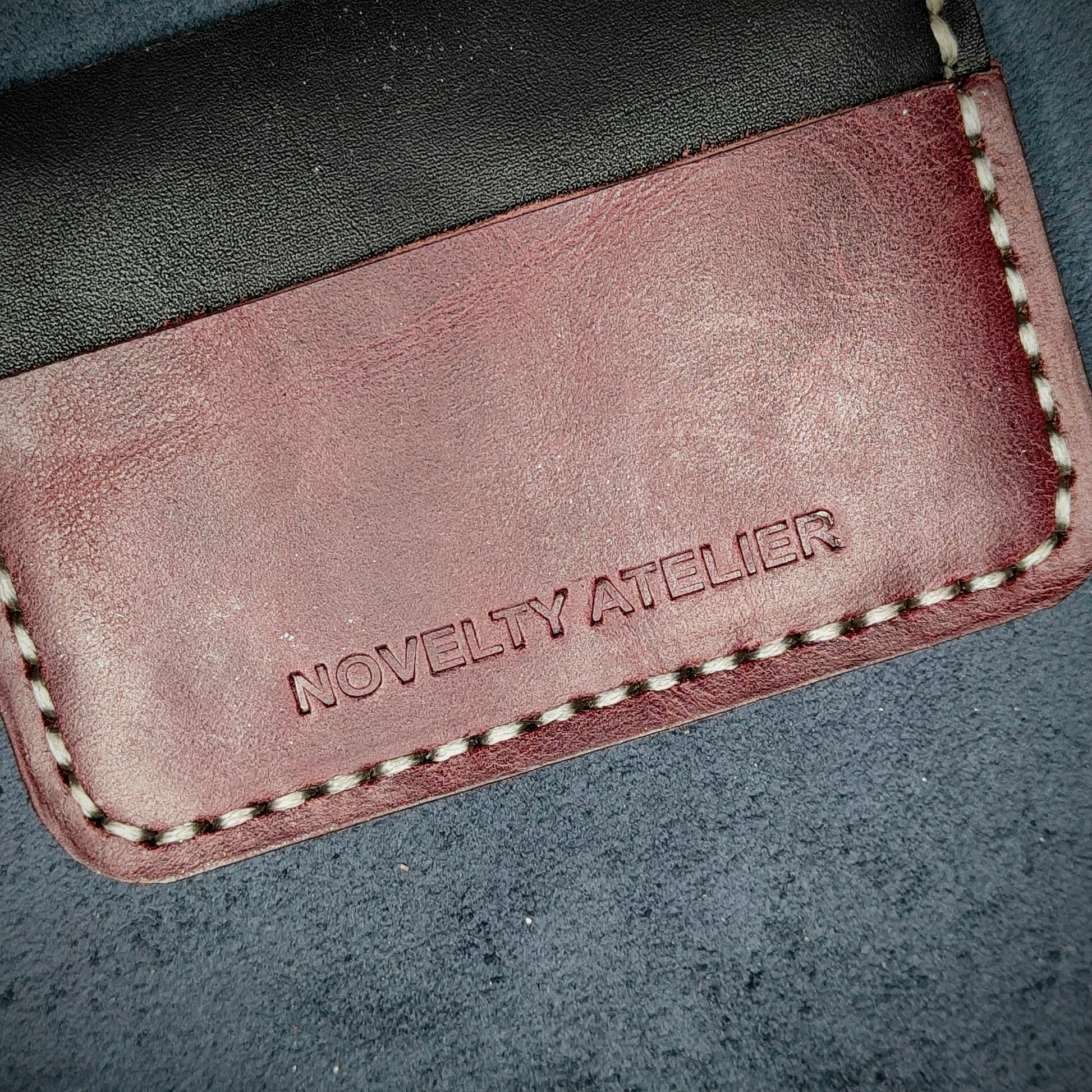 Front view close up shot of the Novelty Atelier brand emboss on The Houdini card holder in horizontal configuration– detailed close-up of premium Black and Purple full-grain leather and contrast off-white hand-stitching . Emphasizes the sharp, elegant logo impression and tactile texture, appealing to luxury EDC and minimalist gear collectors.
