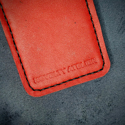 Front view close up shot of the Novelty Atelier brand emboss on The Houdini card holder in vertical configuration– detailed close-up of premium Black and Red full-grain leather and contrast Black hand-stitching . Emphasizes the sharp, elegant logo impression and tactile texture, appealing to luxury EDC and minimalist gear collectors.
