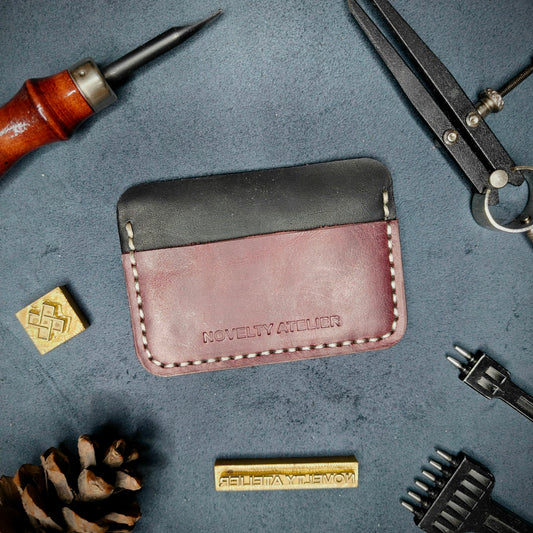 Front view of The Houdini minimalist card holder in horizontal configuration by Novelty Atelier – handcrafted from Black and Purple ethically sourced full-grain leather with contrasting off-white saddle stitching. Features the “Novelty Atelier” brand name cleanly embossed on the front. Perfectly balanced minimalist wallet design tailored for modern EDC enthusiasts who value craftsmanship and brand integrity.
