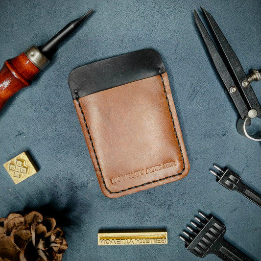 Front view of The Houdini minimalist card holder in vertical configuration by Novelty Atelier – handcrafted from Black and Brown ethically sourced full-grain leather with contrasting Black saddle stitching. Features the “Novelty Atelier” brand name cleanly embossed on the front. Perfectly balanced minimalist wallet design tailored for modern EDC enthusiasts who value craftsmanship and brand integrity.
