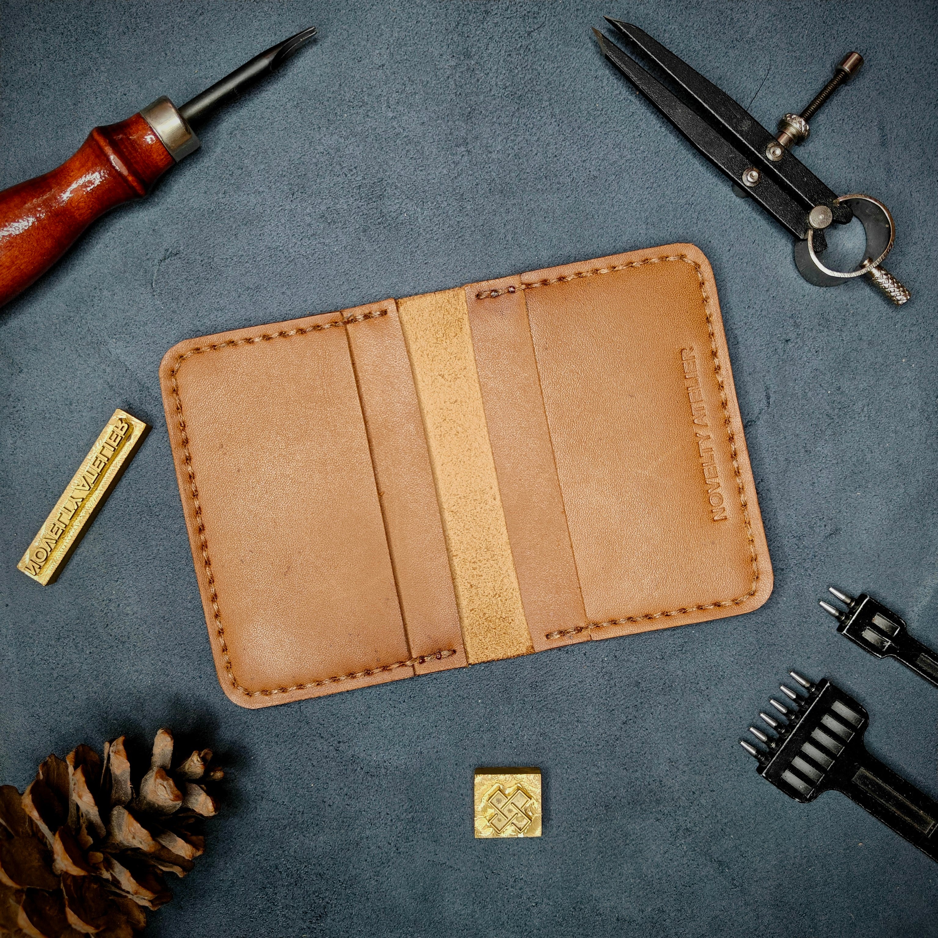 Home page banner of The Copperfield handmade leather card holder by Novelty Atelier open flat view revealing 4 card slots, crafted from ethically sourced Tann full grain leather with brown saddle stitching. Displayed on suede with brass stamp, leatherworking tools, and pine cone for EDC inspired composition. A durable, minimalist wallet for everyday carry.
